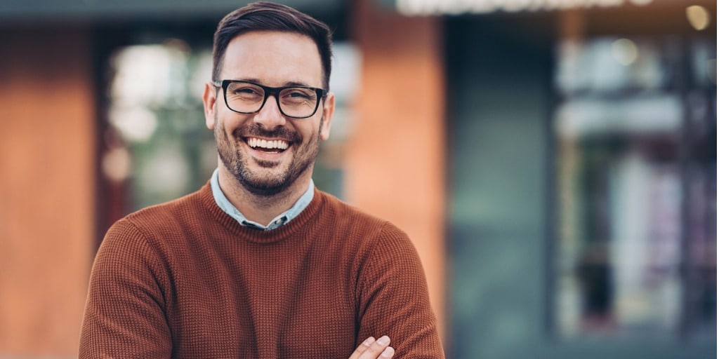 headshot of man in long sleeves folding arms and smiling with glasses, against blurred background of building
