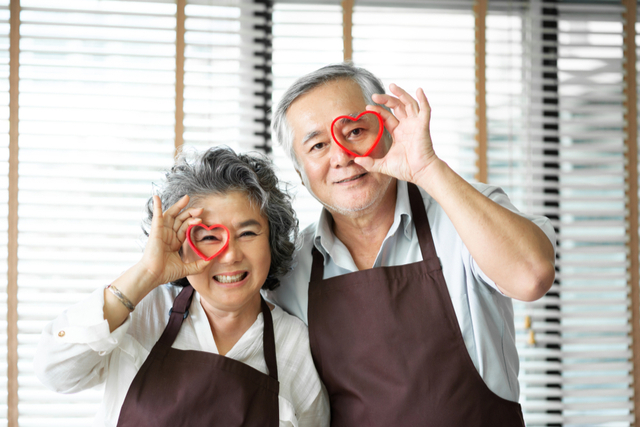 Asian senior couple wear brown aprons are holding red cookies cutters in heart shape over their eyes